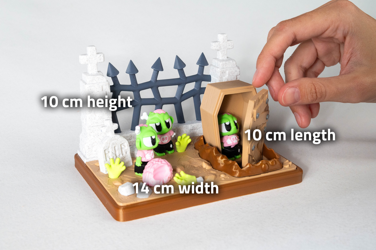 A small toy graveyard scene on a display base with two green cartoon characters, white tombstones, a gray fence, and a brown casket. A human hand is touching the casket, and overlaid text shows dimensions: 10 cm height, 10 cm length, and 14 cm width.